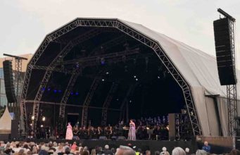 La ópera conquista las calles de ’s-Hertogenbosch con sonido ARA Opera al aire libre con sonido ARA en el corazón de ’s-Hertogenbosch Opera op de Parade reunió a miles de personas en la Parade de ’s-Hertogenbosch, en el sur de Países Bajos, para disfrutar de dos noches de ópera y música clásica al aire libre. El festival, organizado por Opera Zuid, Philzuid y Theater aan de Parade, regresó en 2026 con una edición especial marcada por su 20 aniversario y por una programación concebida para acercar la ópera a públicos muy diversos. La primera jornada estuvo dedicada a la Opera Singalong, una propuesta participativa en la que el público no solo asistió como espectador, sino que formó parte de la experiencia cantando junto a Philzuid, solistas de la International Vocal Competition ’s-Hertogenbosch y Brabant Koor. La segunda noche tuvo como protagonista L’Étoile, la ópera de Emmanuel Chabrier, una obra de carácter satírico, llena de humor y giros inesperados, que transformó la plaza en un gran teatro al aire libre. Para esta edición, la producción técnica estuvo a cargo de Tintaan, con Marty Brugmans como responsable de producción. Jannes Noorman se encargó de la mezcla de FOH, mientras que Miguel Augusto asumió la configuración y optimización del sistema. La solución de sonido se articuló a través de Unlimited Vision & Sound, empresa neerlandesa especializada en soluciones audiovisuales para eventos, con el apoyo de TSL Production y LSP, que aportaron parte del sistema utilizado para la producción. La coordinación local contó además con la implicación del equipo DAS AUDIO Benelux, con Henri y Rob desempeñando un papel clave en el seguimiento del proyecto, la conexión entre los diferentes equipos y el apoyo necesario para que la solución respondiera a las necesidades reales del evento. Reto: claridad, cobertura y control en un entorno abierto El principal reto técnico era trasladar la precisión de una producción operística a un espacio urbano abierto, con un público distribuido en una plaza amplia y sin las condiciones acústicas controladas de un teatro. En un evento de estas características, la cobertura debe ser uniforme, la inteligibilidad vocal resulta crítica y el sistema debe mantener la naturalidad de las voces y de la orquesta sin alterar la dinámica propia de la interpretación. A estas exigencias se sumaba la propia naturaleza del festival. La Opera Singalong requería un sistema capaz de acompañar una experiencia participativa, con miles de asistentes cantando y siguiendo la música en directo, mientras que L’Étoile exigía una reproducción más teatral, con mayor atención al detalle vocal, a los matices orquestales y a la integración entre música, escena y espacio. El entorno de la Parade, en pleno centro de ’s-Hertogenbosch, añadía además una necesidad clave: controlar con precisión la energía proyectada fuera del área de audiencia. En una producción al aire libre y en un contexto urbano, no basta con alcanzar presión sonora suficiente. Es necesario dirigir el sonido hacia el público, reducir la energía no deseada sobre el escenario y limitar la propagación hacia las zonas próximas de la ciudad. Solución: una configuración cardioide ARA para una experiencia operística al aire libre Para responder a estas necesidades, el sistema principal se configuró con 26 MARA, apoyados por 12 LARA-SUB y 10 ARA-P28. La elección de MARA y LARA-SUB resultó especialmente relevante por su diseño cardioide, que permitió mejorar el control de la directividad tanto en medias y bajas frecuencias como en el rango grave del sistema. En un evento al aire libre, esta capacidad de control aporta ventajas muy concretas. Por un lado, reduce la energía proyectada hacia la parte posterior del sistema, disminuyendo el nivel de sonido sobre el escenario y facilitando el trabajo de artistas, músicos y equipo técnico. Por otro, ayuda a contener la dispersión sonora hacia el entorno urbano, algo especialmente importante en una plaza situada en el centro de la ciudad. Los sistemas MARA proporcionaron la base principal de cobertura para la audiencia, con una respuesta diseñada para mantener claridad, definición y consistencia en un formato compacto, pero con la misma filosofía de control de la serie ARA. Su comportamiento permitió cubrir una audiencia amplia con una dispersión horizontal especialmente uniforme, manteniendo una experiencia sonora coherente en diferentes zonas de la plaza. Los LARA-SUB aportaron el refuerzo en bajas frecuencias necesario para sostener la dimensión musical del evento, manteniendo un comportamiento cardioide que contribuyó a reducir la energía no deseada detrás del sistema y a mejorar la limpieza general de la mezcla. Los ARA-P28 completaron la configuración para reforzar zonas específicas de cobertura y mejorar la experiencia del público en áreas donde era necesario un apoyo adicional. La combinación de estos sistemas permitió adaptar la respuesta a las características de la Parade, manteniendo un equilibrio entre presión sonora, inteligibilidad, naturalidad tímbrica y control de la dispersión. La coordinación entre Tintaan, Unlimited Vision & Sound, TSL Production, LSP y el equipo técnico de DAS AUDIO permitió implementar una solución ajustada a las necesidades reales del evento. El trabajo de Miguel Augusto como técnico de sistema fue clave para optimizar la configuración, ajustar la cobertura y garantizar que el sistema respondiera de forma precisa tanto durante la Opera Singalong como en la representación de L’Étoile. Conclusión: “Lo que más nos sorprendió fue la dispersión horizontal del sistema” La respuesta del sistema superó las expectativas del equipo técnico y de producción, que destacó especialmente la claridad, el control y la naturalidad conseguidos en una plaza abierta y en pleno entorno urbano. “Lo que más nos sorprendió fue la dispersión horizontal del sistema. En una plaza abierta, con una audiencia distribuida de forma muy amplia, necesitábamos una cobertura consistente, natural y bien controlada. El sistema consiguió mantener esa sensación de uniformidad sin perder definición ni generar más energía de la necesaria fuera del área de audiencia”, señaló Marty Brugmans, responsable de producción de Tintaan. Desde FOH, Jannes Noorman también valoró la respuesta del sistema durante las dos jornadas del festival. “Para ópera al aire libre, la inteligibilidad y la dinámica son fundamentales. El sistema ofreció una imagen muy estable, con voces claras, una respuesta musical muy natural y un escenario más limpio de lo habitual en este tipo de producciones”, explicó. La edición 2026 de Opera op de Parade demostró cómo una configuración basada en la serie ARA puede trasladar la precisión de una producción escénica a un espacio urbano abierto, manteniendo el equilibrio entre cobertura, control, reducción de energía no deseada y calidad sonora. La colaboración entre Theater aan de Parade, Unlimited Vision & Sound, Tintaan, TSL Production, LSP, Rob Pigmans y el equipo DAS AUDIO Benelux hizo posible una experiencia en la que la ópera se vivió de forma cercana, participativa y con un alto nivel técnico. Créditos del proyecto Evento: Opera op de Parade Producción: Tintaan Responsable de producción: Marty Brugmans FOH: Jannes Noorman Técnico de sistema: Miguel Augusto Sistema: DAS AUDIO Rental partners: TSL Production y LSP Coordinación local: DAS AUDIO Benelux, Henri y Rob Con la colaboración de: Theater aan de Parade, Unlimited Vision & Sound y Rob Pigmans
