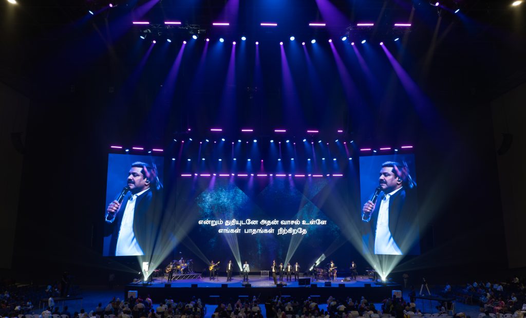 El espacio, con capacidad para 25.000 personas, incorpora una consola grandMA3 light para gestionar alrededor de 180 luminarias en producciones en directo, servicios religiosos y retransmisiones audiovisuales.

El New Life Assembly of God (NLAG) Worship Center, situado en Chennai, al sur de la India, ha incorporado una consola grandMA3 light de MA Lighting como sistema principal de control de iluminación de sus nuevas instalaciones. El recinto multifuncional, con capacidad para 25.000 asistentes, está diseñado para acoger servicios religiosos, conciertos, conferencias y producciones audiovisuales de gran formato.

El proyecto, dirigido por el pastor ejecutivo Benjamin Mohan, planteaba la necesidad de disponer de una plataforma de control flexible, fiable y preparada para futuras ampliaciones. Entre los requisitos principales se encontraban la posibilidad de almacenar y recuperar escenas y programaciones de forma consistente, así como gestionar diferentes configuraciones de iluminación en función de las características de cada evento.

La instalación cuenta con aproximadamente 180 luminarias distribuidas en cinco estructuras truss, controladas mediante múltiples universos DMX. La infraestructura combina la consola grandMA3 light con nodos de red de terceros y 28 distribuidores DMX, formando un sistema que centraliza la programación, la reproducción de secuencias, la ejecución de cues y los eventos sincronizados mediante código de tiempo.

Un sistema de control adaptable a diferentes producciones

La configuración diáfana del recinto permite modificar la disposición del escenario y del público según las necesidades de cada celebración o producción. Esta versatilidad exige un sistema de iluminación capaz de adaptarse a distintos equipos técnicos, operadores y formatos de espectáculo.

Según explica Theophorus Samuel, director de iluminación de NLAG, una de las características determinantes para seleccionar grandMA3 fue su capacidad de trabajo multiusuario. Esta funcionalidad permite que varios operadores y voluntarios trabajen simultáneamente sobre un mismo archivo de espectáculo, facilitando la coordinación durante la preparación y ejecución de los eventos.

La arquitectura de red se ha diseñado teniendo en cuenta la escalabilidad, la redundancia y la continuidad operativa, tanto en los servicios habituales como en las producciones de mayor complejidad.

En cuanto a la distribución de las luminarias, la primera estructura truss incorpora principalmente proyectores de perfil destinados a la iluminación principal del escenario y a aplicaciones específicas. Las cuatro restantes combinan cabezas móviles wash, spot y luminarias blinder.

Samuel señala que el objetivo durante los servicios habituales es crear un entorno inmersivo y confortable, evitando efectos excesivos que puedan distraer a los asistentes. La iluminación debe acompañar el desarrollo de cada celebración y contribuir a la experiencia visual sin adquirir un protagonismo innecesario.

Esta filosofía también responde a la creciente importancia de las retransmisiones televisivas y el streaming en los centros religiosos del sur de la India, donde la iluminación escénica desempeña un papel cada vez más relevante en la calidad de las producciones audiovisuales.

Programación colaborativa y visualización 3D

La elección de grandMA3 supuso un cambio respecto a los sistemas de control utilizados anteriormente por NLAG en sus otras instalaciones de Chennai.

Durante la fase de diseño del nuevo recinto, las consultoras Salas O'Brien y HouseRight desarrollaron los conceptos audiovisuales, acústicos y los diseños esquemáticos en colaboración con el estudio internacional de arquitectura Populous y el equipo de NLAG.

HouseRight propuso inicialmente la incorporación de grandMA3, una recomendación que posteriormente fue evaluada por Benjamin Mohan y Theophorus Samuel. En el desarrollo y la implementación del proyecto también participaron Franco Zaghini, director de desarrollo de negocio de MA Lighting, el diseñador de iluminación local Viju y Manuel Raj, director de medios de NLAG.

Uno de los aspectos más valorados por el equipo técnico es la integración de grandMA3 con los formatos GDTF y MVR, utilizados para la gestión de perfiles de luminarias, las copias de seguridad y la preparación de entornos de previsualización tridimensional.

Esta compatibilidad facilita la correspondencia entre la programación y la representación virtual de los equipos, además de simplificar la preparación de diferentes configuraciones de espectáculo.

Samuel destaca igualmente las herramientas de programación Recipes, Cues y Sequences, que permiten realizar modificaciones sobre múltiples luminarias y actualizar contenidos sin necesidad de reconstruir por completo los espectáculos. Estas funciones resultan especialmente útiles en un recinto que trabaja con diferentes equipos de producción y operadores voluntarios.

Los recursos de ayuda integrados en grandMA3 también contribuyen a la formación de nuevos usuarios y a la resolución de incidencias durante la operación.

Soporte técnico y puesta en marcha

La consola grandMA3 light fue suministrada por Hi-Tech Audio & Image, distribuidor oficial de MA Lighting en India, que también proporcionó asistencia técnica durante el proyecto.

Con el nuevo sistema en funcionamiento, NLAG dispone de una infraestructura de control preparada para gestionar tanto sus servicios semanales como conciertos, encuentros juveniles, representaciones teatrales y otros eventos de gran formato.

Para Benjamin Mohan, el resultado más importante es conseguir que todos los elementos técnicos funcionen de manera coordinada y que la tecnología contribuya a la experiencia de los asistentes sin interferir en el desarrollo de las celebraciones.